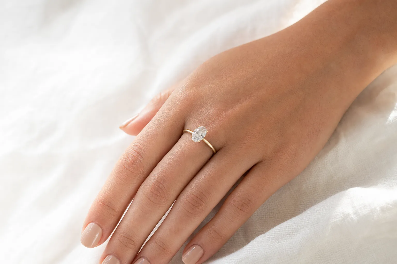 Solitär-Verlobungsring mit ovalem Labordiamant in Gelbgold auf einer Hand, ruhende Position auf weissem Leinen