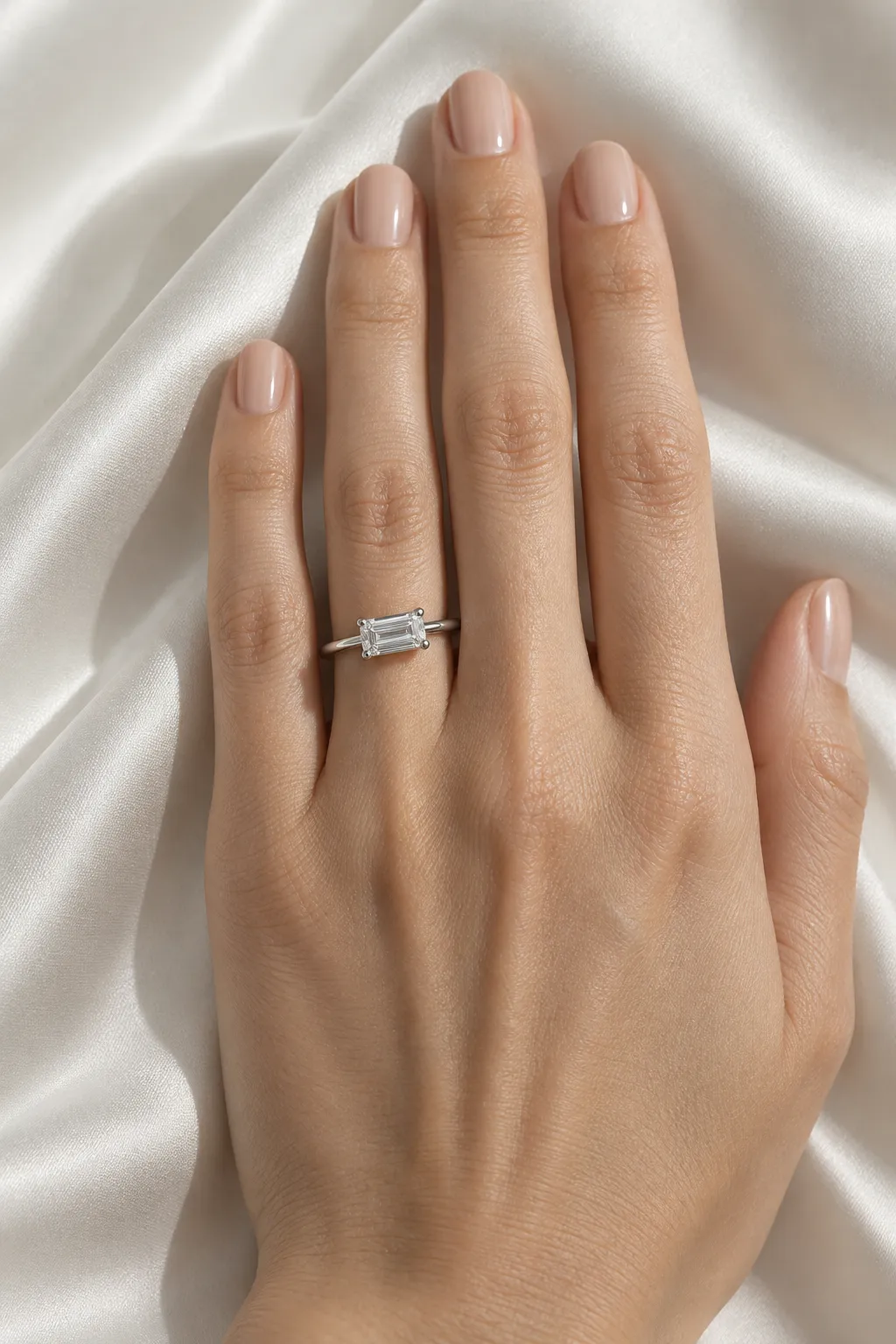Hand auf weisser Seide mit East-West-Verlobungsring in Weissgold, der Emerald-Cut-Diamant liegt quer über dem Ringfinger