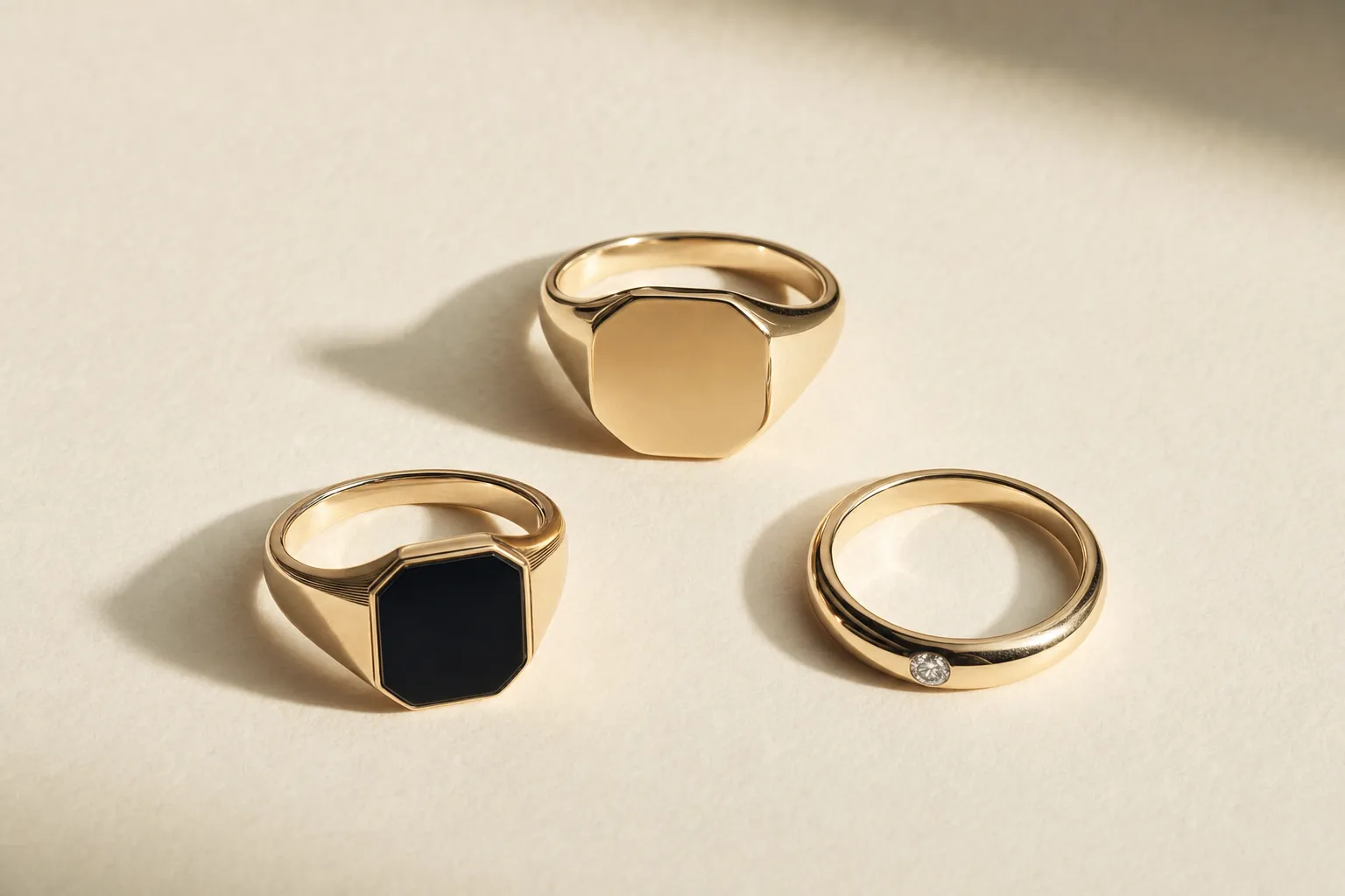 Drei Herrenringe auf warmem Weiss: ein Siegelring, ein schwarzer Onyx-Pinky-Ring in Gold, ein Ring mit einem kleinen Diamanten